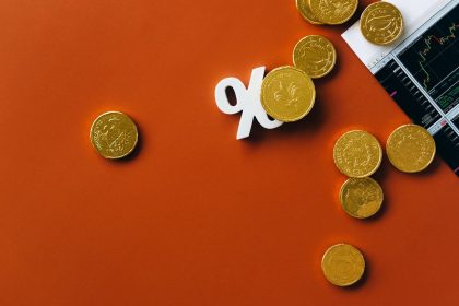 Moedas douradas ao lado de um gráfico de mercado e do símbolo de porcentagem sobre fundo laranja.