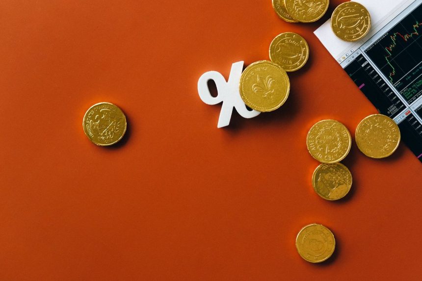 Moedas douradas ao lado de um gráfico de mercado e do símbolo de porcentagem sobre fundo laranja.