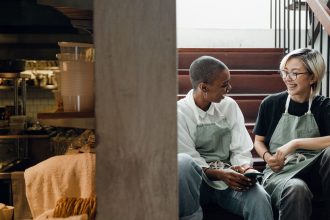 Duas trabalhadoras usando aventais descansam e conversam sentadas em uma escada próxima a uma cozinha.